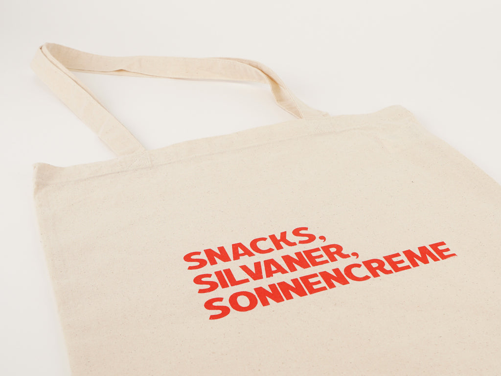 Detailaufnahme einer Baumwolltasche mit dem roten Aufdruck „SNACKS, SILVANER, SONNENCREME" auf naturfarbenem Stoff